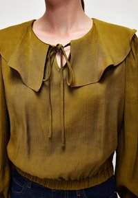 Oliven grøn bluse med en stor, flagrende krave, bindebånd ved halsen og elastisk talje. Stoffet har en subtil glans og struktureret overflade.