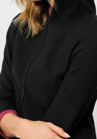 Sweat à capuche noir texturé avec un col montant, présentant un détail de poignet rose contrastant. Le matériau semble doux et tricoté.