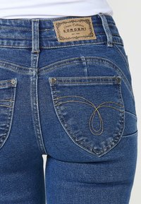 Los vaqueros de mezclilla azul tienen un lavado medio, con una etiqueta de cuero en la cintura y costuras decorativas en los bolsillos traseros.