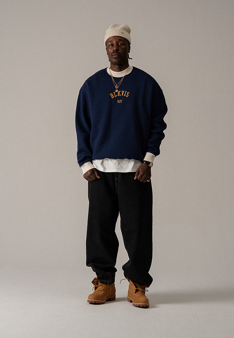 Tengerészkék pulóver sárga felirattal, fehér inggel rétegezve. Fekete bő, laza farmernadrág és barna csizma egészíti ki az öltözéket. Semleges háttér.