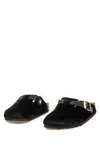 Zuecos de felpa negra sin cordones con correas ajustables negras y suelas planas marrones, presentados sobre un fondo blanco.