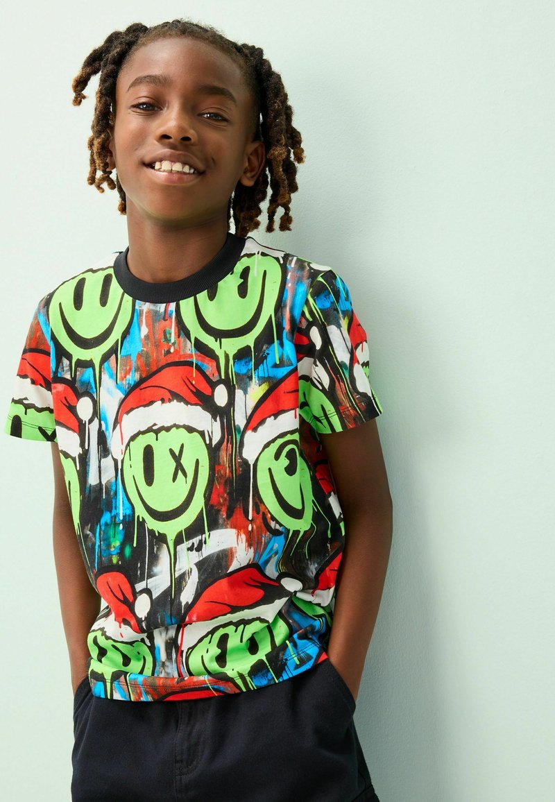 Bambino sorridente con dreadlocks che indossa una maglietta colorata con faccine verdi sorridenti con cappelli di Babbo Natale e pantaloncini neri, in piedi contro un muro chiaro.
