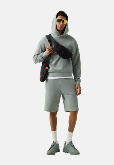 Mann trägt grauen Hoodie und Shorts, übergroße Sonnenbrille, weiße Socken, grüne Turnschuhe und zwei schwarze Umhängetaschen auf grauem Hintergrund.