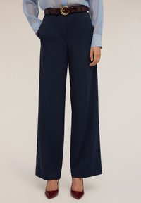 Pantaloni a gamba larga blu navy realizzati in tessuto morbido, con vita alta e tasche laterali, abbinati a una cintura marrone con dettaglio della fibbia dorata.