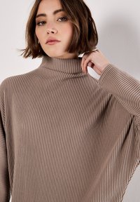 Maglione a collo alto a coste marrone con maniche lunghe, caratterizzato da una vestibilità ampia e un tessuto testurizzato, che mette in risalto linee verticali e motivi sottili.