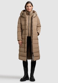 Dlouhý, prošívaný kabát v světle béžové barvě s kapucí, vybavený zipovým zapínáním, bočními kapsami a značkovou visačkou. Nositelný přes odpovídající vnitřní bundu.