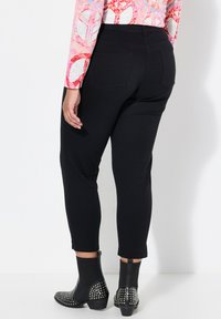 Schwarze Stretchhose mit schlanker Passform, mittelhoher Taille und Taschen hinten. Kombiniert mit genagelten schwarzen Stiefeletten. Die Textur wirkt weich.