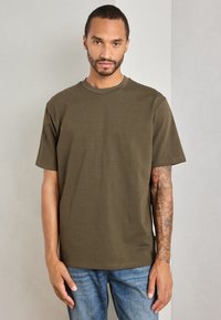 Bărbat cu păr scurt și barbă, îmbrăcat într-un tricou verde măsliniu și blugi, stând în fața unui zid simplu de culoare deschisă, arătând un braț drept tatuat.
