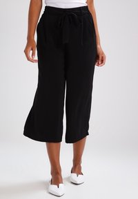 Svarta korta byxor med dragsko i midjan, lättviktsmaterial och vida ben, kombinerade med vita skor med spetsig tå.