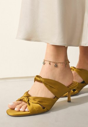 Füße in senfgelben Satin-Sandalen mit Absätzen, verdrehten Riemen und einem goldenen Fußkettchen, gezeigt mit einem beigen Rocksaum.