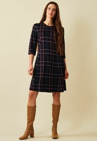 En rutig klänning i mörk marinblå och burgundy med avslappnad passform, trekvartslånga ärmar och utsvängd nederdel. Matchad med bruna knähöga stövlar.