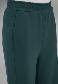 Pantalons de sport vert foncé en tissu doux et léger, avec une taille élastique et deux poches latérales. Texture lisse.