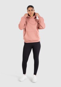 Haut à capuche en rose surdimensionné avec une poche avant et des cordons ajustables, associé à des leggings noirs et des chaussures de sport blanches.