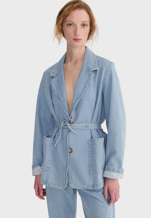 Młoda kobieta o bladej karnacji i rudych włosach ubrana w jasnoniebieską kurtkę dżinsową i pasujące spodnie, stojąca na białym tle.