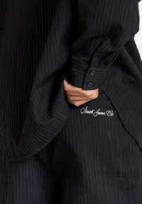 Zwarte gebreide shirt met verticale ribbels, knoopmanchetten en geborduurd logo aan de zijkant, dat een losse pasvorm en zachte stof laat zien.