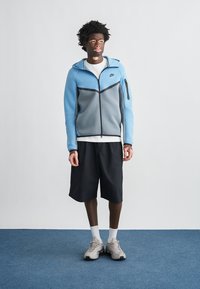 Μπλε και γκρι ζακέτα με φερμουάρ και μαύρες λεπτομέρειες, φορεμένη πάνω από λευκό πουκάμισο. Μαύρες βερμούδες και αθλητικά παπούτσια συμπληρώνουν την εμφάνιση. Απλό φόντο.