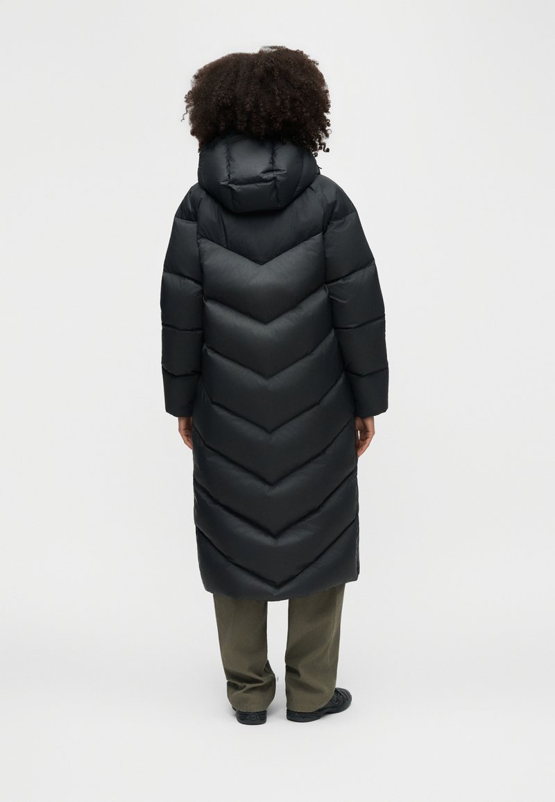 Person mit lockigem Haar, der einen langen schwarzen Steppmantel mit Chevron-Steppung, olivgrüne Hose und schwarze Schuhe trägt und von hinten zu sehen ist.