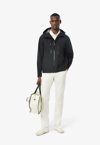 Giacca nera impermeabile con tasche con zip, camicia bianca, pantaloni chiari e borsa crema con dettagli neri e zip.