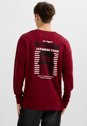 Joven con una camiseta de manga larga color granate con un gráfico en blanco y negro de comida japonesa y texto en la espalda.