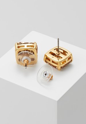 Boucles d'oreilles carrées dorées avec paillettes incrustées, tiges en métal et attaches papillon en plastique transparent sur une surface blanche.