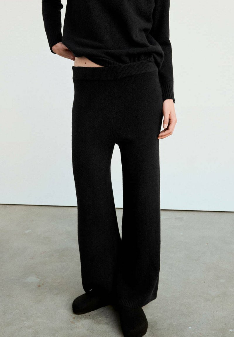 Personne portant un pantalon en maille noire ample et un pull assorti, debout sur un sol en béton contre un mur blanc.