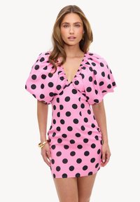 Femme portant une courte robe rose avec de gros pois noirs, avec un profond décolleté en V et des manches bouffantes surdimensionnées.