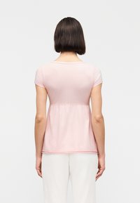 Γυναίκα με κοντά σκούρα μαλλιά που φοράει ανοιχτό ροζ μπλούζα με κοντά μανίκια και λευκό παντελόνι, εμφανιζόμενη από πίσω με φόντο λευκό.