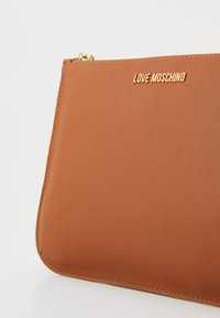 Borsa a tracolla in pelle marrone con una texture liscia e un logo dorato, chiusura con zip e bordi cuciti. Design compatto e rettangolare.