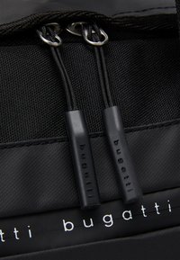 Black fabric backpack close-up with two zipper pulls, textured material, and 'bugatti' branding in white on the front panel.