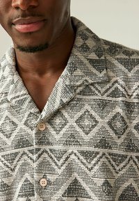 Camisa de manga corta en tejido plano con un patrón geométrico en beige y negro, con un cuello de muesca y botones crema.