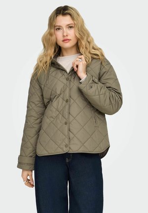 Žena s valovitom plavom kosom, odjevena u kaki zeleno prošivenu jaknu i tamnoplave traperice, drži ovratnik jakne desnom rukom.