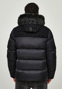 Mand iført en sort puffjakke med en hætte beklædt med pels og geometrisk mønster på den øverste ryg, stående mod en ensfarvet baggrund.