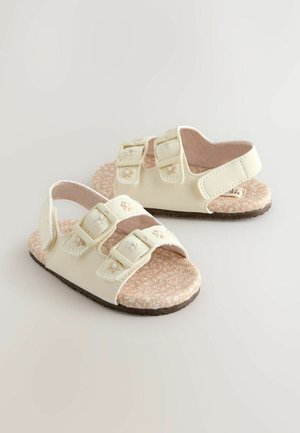 Svijetlo bež sandale s dva podesiva remena sa kopčama, teksturiranim uloškom i potplatom s uzorkom nalik pluti. Imaju cvjetni vezeni ukras na remenima.