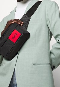 Sac en bandoulière en tissu noir avec une patch rectangulaire rouge portant le logo "HUGO", associé à une veste de costume texturée vert clair.