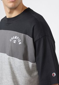 Hombre vestido con una camiseta Champion de bloques de color negro, gris oscuro y gris claro, con el logo en el pecho y en la manga, contra un fondo blanco.