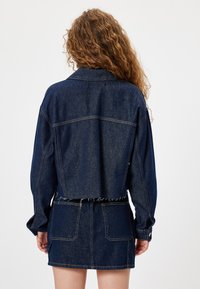 Giacca di jeans blu scuro con un design cropped, maniche lunghe e dettagli a orlo sfrangiato, abbinata a una minigonna in jeans coordinata.