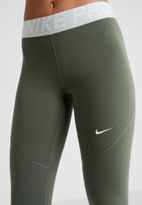 Nike Pro leggings korkealla vyötäröllä oliivinvihreänä, valmistettu sileästä, joustavasta kankaasta, jossa on valkoinen logo ja vyötärönauhan yksityiskohtia.