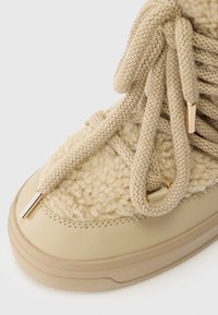 Calzature beige con una tomaia texturizzata simile alla lana, lacci piatti e dettagli dorati sui passanti e sugli aglet. Suola in gomma liscia.