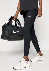 Un legging noir et un sac de sport noir Nike sont tenus dans une main. Les chaussures de sport sont blanches avec des accents noirs. Les logos Nike sont visibles.
