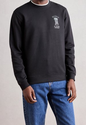 Czarna bluza z okrągłym dekoltem z białym napisem „Scotch & Soda Amsterdam” i grafiką kolumny na piersi, noszona z niebieskimi dżinsami przez stojącą osobę.