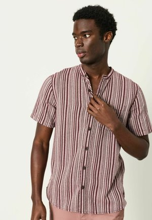 Mann mit kurzen lockigen Haaren, der ein kurzärmeliges, vertikal gestreiftes Hemd in Brauntönen trägt und den Kragen anpasst, während er nach links blickt.