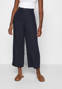 Breda byxor i mörk marinblå linne med hög midja, framknäppning och en avslappnad passform. Matchas med bruna platta sandaler.