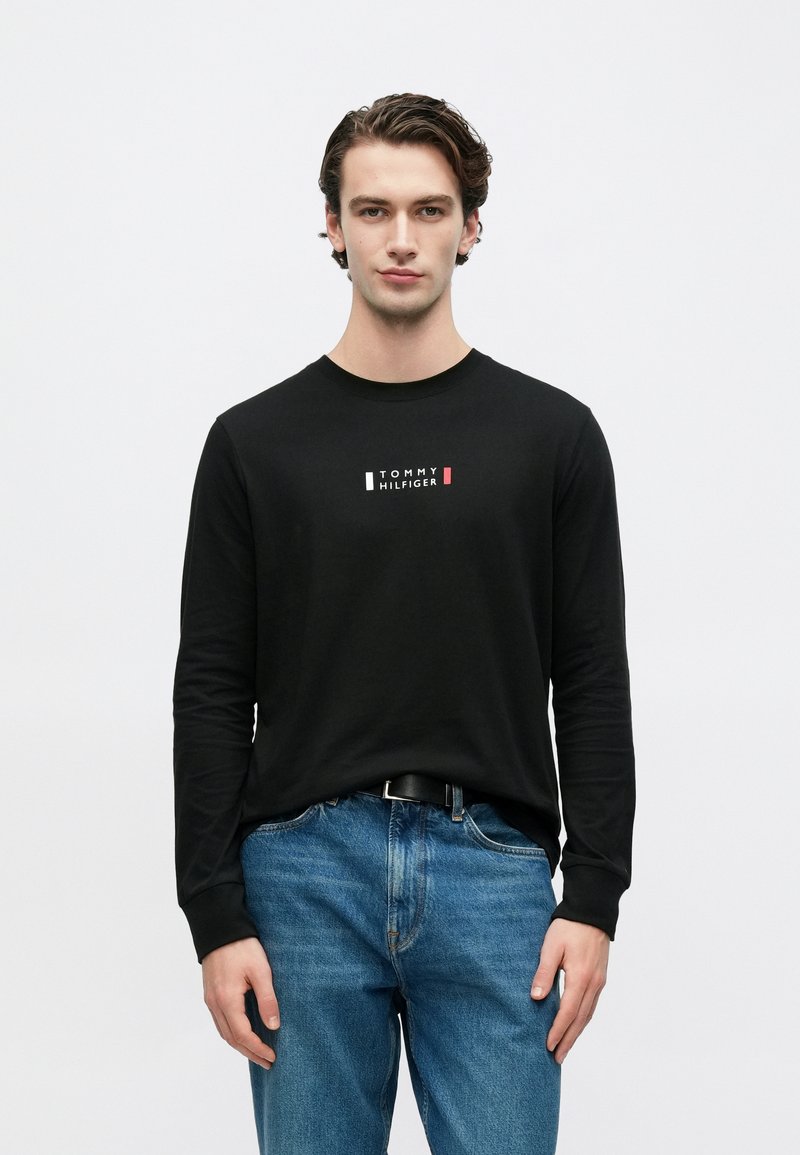 Black long-sleeve t-shirt with a small "TOMMY HILFIGER" logo in white, red, and green. Paired with blue denim jeans.