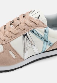 Tennised, millel on beež nubukist ja valge tekstiili ülemine osa, peegeldavad hõbedased detailid ja beežid paelad. Küljel olev logo kannab kirja "Armani Exchange."