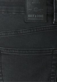 Gros plan sur la poche arrière d'un jean en denim noir avec une étiquette cousue portant la mention "ONLY & SONS FABRICANTS DE JEANS PASSIONNÉS."
