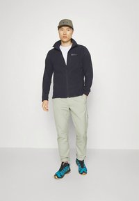 Black fleece zip-up jacket, light green pants, and blue/black sneakers. Model wears a gray cap, standing against a light background.