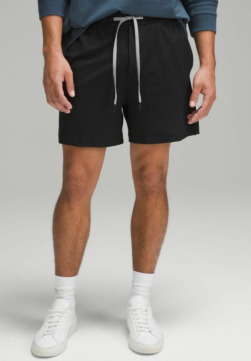 Shorts de sport noirs avec une taille élastique et un cordon de serrage gris. Portés avec des baskets blanches et des chaussettes de longueur moyenne. Texture de tissu lisse.
