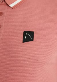 Polo à manches courtes en tissu texturé rose, avec une patte carrée noire ornée d'un motif géométrique blanc et un col à rayures blanches.