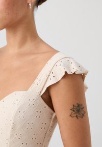 White dress with intricate embroidered pattern, shoulder ruffles, and structured bodice. Visible floral tattoo on the upper arm.