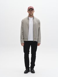 Jachetă gri deschis cu fermoar peste un tricou alb, asortată cu pantaloni negri și pantofi negri. O șapcă bordo cu text alb completează ținuta.